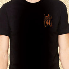 A person wears Jeff Granitos 44 Mai Tai Unisex Tee, featuring a small brown cocktail glass and 44 on a black shirt with decorative elements and a textured background.