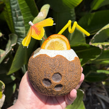 A hand holds a limited-edition Kokomo Coconut Ceramic Tiki Mug, featuring a straw and a yellow paper umbrella, with an orange slice inside. The mug is set against tropical green leaves, evoking island vibes—ideal for those pre-ordering mugs to savor tropical drinks.