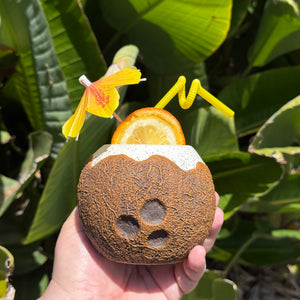 A hand holds the Kokomo Coconut Ceramic Tiki Mug, featuring a textured coconut shape. Its garnished with an orange slice, a decorative yellow flower, and a curly straw. Green leaves enhance the tropical feel of this limited-time pre-order experience.