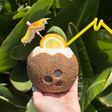 A hand elegantly holds a Kokomo Coconut, ceramic tiki mug, adorned with an umbrella, orange slice, and curly straw. Lush green leaves whisper of adventures and limited-time treasures awaiting just beyond the horizon.