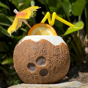 The Kokomo Coconut Ceramic Tiki Mug, featuring a textured brown surface and adorned with a yellow umbrella, orange slice, and bendy straw, sits on a rock. Available for a limited time pre-order, it awaits amidst the backdrop of blurred green leaves.