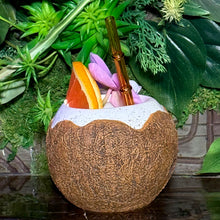 The Kokomo Coconut Ceramic Tiki Mug features a tropical design with a coconut shell appearance, frothy white rim, orange slice garnish, pink flower, and brown straw against a greenery backdrop. Enjoy this exclusive drinkware delight while it lasts—limited quantity available for pre-order.