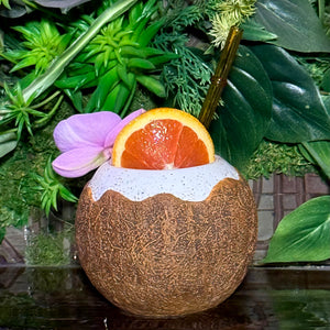 The Kokomo Coconut Ceramic Tiki Mug, available for a limited-time pre-order, is a decorative mug styled like a coconut shell. It features a slice of orange and purple flower as garnish and comes with a straw—perfect for those seeking unique tropical experiences against lush green foliage.