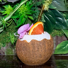 The Kokomo Coconut Ceramic Tiki Mug, available for limited-time pre-order, is coconut-shaped, comes with a colorful drink garnished with an orange slice and pink flower, and features a stylish gold straw. Its set against lush green foliage for a perfect tropical vibe.