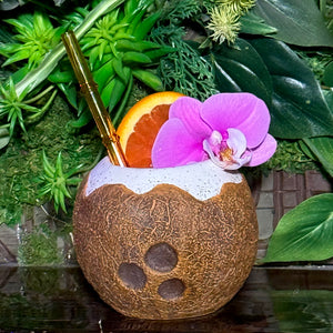 Sip a tropical cocktail served in a coconut-like ceramic mug, adorned with a pink orchid and orange slice, featuring a gold straw. Enjoy this exotic experience with our Kokomo Coconut - Ceramic Tiki Mug, available for limited-time pre-orders.