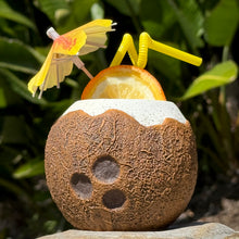 The Kokomo Coconut Ceramic Tiki Mug features a textured coconut design with a white rim, adorned with an orange slice and yellow cocktail umbrella. Two yellow straws complete the vibe. Nestled against lush green foliage, this limited edition mug is available for pre-order now!.