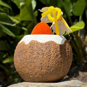 The Kokomo Coconut Ceramic Tiki Mug features a frothy white top and cradles a bright orange drink, topped with a yellow straw and paper umbrella. Available for limited-time pre-order, it sits against blurred green leaves.