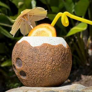 The Kokomo Coconut Ceramic Tiki Mug, available for limited-time pre-order, is a coconut-shaped cup with a rough brown exterior and smooth white top. It features a yellow spiral straw, an orange slice, and a petite paper umbrella, making it perfect against lush green foliage in your collection.