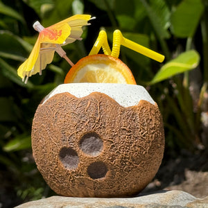 The Kokomo Coconut Ceramic Tiki Mug features a tropical coconut drink with an orange slice and yellow umbrella, set on stone with lush green leaves. Two yellow straws complete the look. Pre-order now—limited quantities available for your collection!.