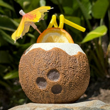 The Kokomo Coconut Ceramic Tiki Mug, available for a limited-time pre-order, sits on a rock filled with a drink, garnished with an orange slice, pink paper umbrella, and yellow straws. Lush foliage in the background suggests a tropical setting where you can almost hear the waves.